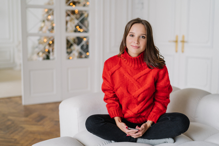 Photo of cute European woman has dark hair, dressed in domestic clothes, spends winter weekend at home, sits in lotus pose on comfortable sofa, uses moden cell phone for online communicationの写真素材
