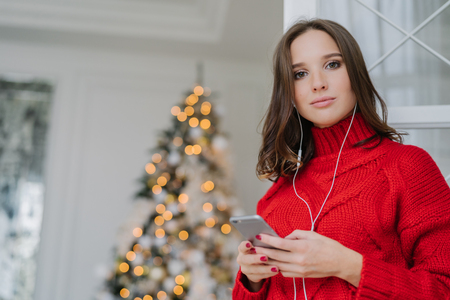 Photo of good looking female with dark hair, dressed in red knitted sweater, surfes social networks on mobile phone, listens music with earphones, admires decorated Christams tree. Leisure conceptの写真素材