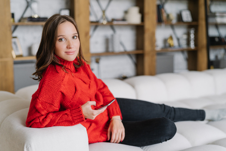 Photo of attractive female model in knitted red sweater, messages online at home, poses on couch in living room, uses modern cell phone and wireless internet connection, looks with appealing lookの写真素材