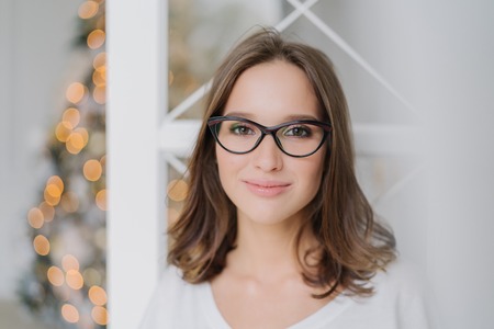 Close up shot of good looking young female model wears spectacles for good vision, looks happily at camera, has pleased expression, charming smile, poses against decorated Christmas tree indoorの写真素材