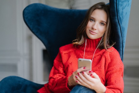 Photo of charming female sits in comfortable armchair, dressed in sweater, listens audio book, connected to earphones, uses modern cell phone and high speed internet connection. Leisure conceptの写真素材
