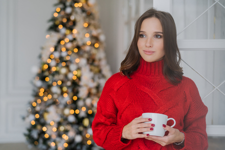 Photo of thoughtful female model wears red sweater, drinks tea, has dark hair, contemplates about something, stands in spaciouss room near decorated new Year tree with lights. Holiday conceptの写真素材