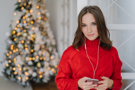 Photo of cute young woman with dark hair, dressed in knitted red sweater, holds smart phone in hands, listens radio online, connected to wifi at home, pose near beautiful New Year tree with lightsの写真素材