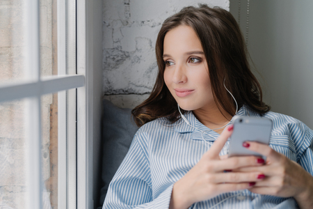 Thoughtful brunette woman listens audio book, holds modern smart phone in hands, enjoys perfect sound in earphones looks pensively at window dressed in casual clothes. Female listens music in playlistの写真素材