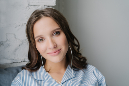 Close up shot of pretty young woman with pleasant appearance listens music with earphones, enjoys spare time at home, poses indoor, has charming look, connected to unrecognizable modern deviceの写真素材