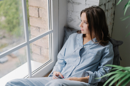 Horizontal shot of thoughtful young woman sits on windowsill, dressed in casual nightwear, uses smart phone and earphones, listens calm music while looks thoughtfully in window, enjoys home timeの写真素材