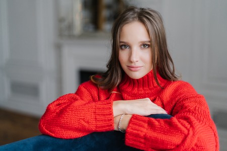 Photo of young good lookng brunette woman enjoys day off at home, has serious look at camera, wears warm loose red sweater, poses at home against blurred background. Coziness and rest conceptの写真素材