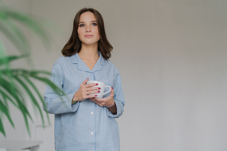 Horizontal shot of pretty woman dressed in oversized striped shit, has attractive loo, holds cup of coffee or latte, stands indoor with blank copy space on left for your advertising content or textの写真素材