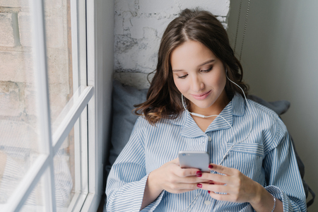 Charming woman enjoys romantic playlist, perfect sound in earphones, holds modern smart phone, dials number of boyfriend, sits on window sill, connected to wireless internet. Rest and home conceptの写真素材