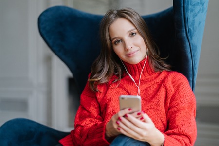 People and technology concept. Satisfied female meloman enjoys favourite music, listens audio record, dressed in red sweater, surfes in playlist, sits in armchair in living room, has spare timeの写真素材