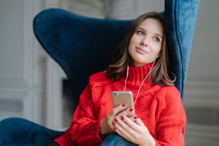 Pensive cute female feels relaxed, listens pleasant music in playlist, holds cellular, looks with dreamy expression aside, poses in comfortable armchair. People, modern technologies and cozinessの写真素材
