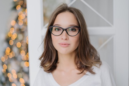 Headshot of lovely young female model in transparent glasses, has dark hair, dressed in casual clothes, enjoys domestic atmosphere, poses against New Year tree. People and lifestyle concept.の写真素材