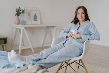 Indoor shot of relaxed female dressed in pyjamas, sits on chair, keeps legs on bed, drinks hot coffee, poses against domestic interior, has appealing look. People, rest and relaxation conceptの写真素材