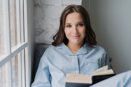 Candid shot of pretty young model sits near window, reads interesting book, looks with satisfied expression at camera, has spare time during weekend. People, leisure, hobby and relaxation conceptの写真素材