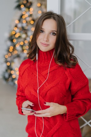 Vertical shot of pleasant looking brunette female model dressed in loose warm sweater, carries smart phone, uses earphones for listening music, poses in domestic interior near decorated fir treeの写真素材
