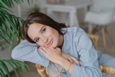 Image of good looking young female model with dark hair, sits on wooden chair, has relaxed facial expression, has charming smile, enjoys domestic calm atmosphere. Beauty and emotions conceptの写真素材