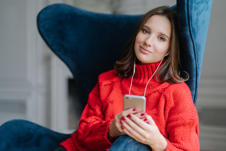 Pretty female teenager enjoys free time and popular music in earphones, holds modern smart phone, dressed in red knitted sweater, sits in comfortable armchair, uses application, checks new updatesの写真素材