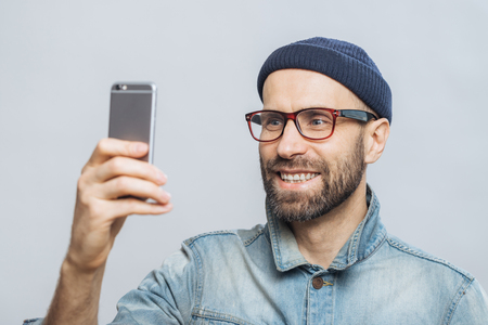 Portrait of happy middle aged smiling male wears denim jacket, hat and glasses, holds smart phone for making selfie, isolated over white background. People, technology and lifestyle concept.の写真素材