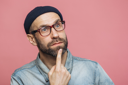 Close up shot of thoughtful bearded male with blue eyes being deep in thoughts, looks pensively into distance, ponders about something, isolated over pink background. Attractive man poses indoorの写真素材