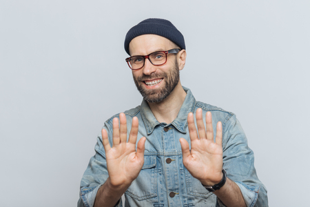 Happy unshaven young male with attractive look shows palms as denies something or demonstrates his refusal, has happy expression, wears stylish black hat, denim jacket and glasses, poses indoorの写真素材