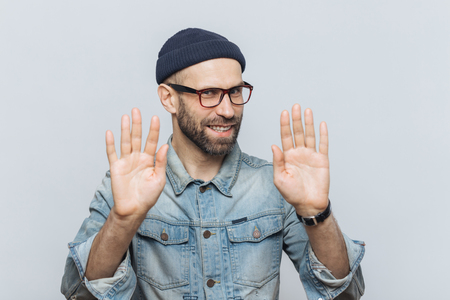 Don`t bother me! Cheerful male with pleasant appearance shows stop sign, asks not to speak with him, wears fashionable hat and glasses, isolated over grey studio background. People and denial conceptの写真素材