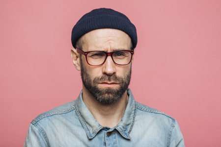 Unhappy discontent unshaven male looks with grumpy expression into camera, wears glasses black hat and stylish denim shirt, isolated over pink background. Displeased middle aged man poses indoorの写真素材