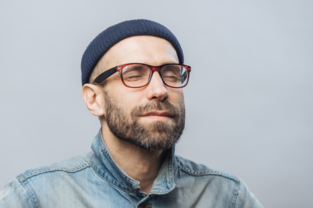 Portrait of relaxed unshaven male with closed eyes wears glasses, keeps eyes closed, enjoys calm atmosphere, isolated over white background. People, relaxation and facial expressions conceptの写真素材