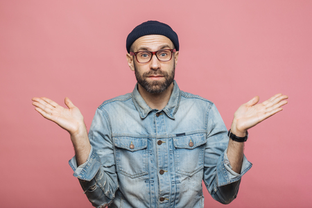Photo of attractive bearded male wears fashionable clothing, shrugs shoulders in bewilderment, makes serious decision, isolated over pink background. Confused hesitant unshaven stylish manの写真素材