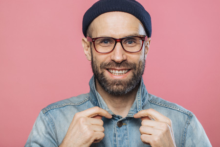 Horizontal shot of pleasant looking smiling bearded male wears glasses and denim jacket, advertizes new fashionable clothes, has happy expression, isolated over pink background. Positive emotionsの写真素材
