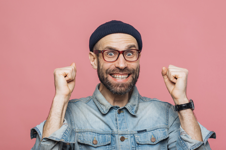 Joyful man with thick beard and mustache clenches fists and looks joyfully at camera, celebrates his victory, has happy expression, isolated over pink background. People, emotions and successの写真素材