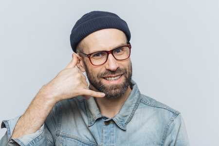 Call me please! Happy handsome man holds hand near ear, imitates mobile phone conversation, smiles joyfully, waits for call from someone, isolated over grey studio background. Communication conceptの写真素材