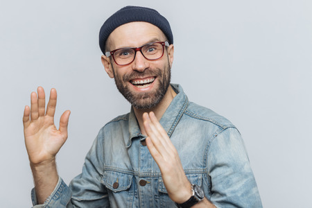 Portrait of happy middle aged man has fun indoor, raises hands, has pleased expression, wears glasses, hat and denim shirt, isolated over white background. People, positive emotions and feelingsの写真素材