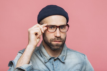 Handsome bearded man with serious expression wears spectacles and glasses, dressed in denim fashionable shirt, isolated over pink background. People, facial expressions and emotions concept.の写真素材
