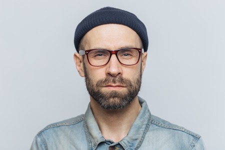 Photo of intelligent confident stylish man with dark thick beard and mustache, looks seriously into camera, poses against grey studio background, wears spectacles and hat. Facial expressions conceptの写真素材