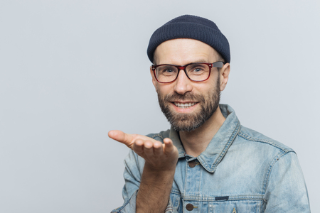Glad bearded male in trendy eyewear expresses his love, shows air kiss, has cheerful expression, isolated over grey background. Blue eyed middle aged man in stylish clothing, gestures indoorの写真素材