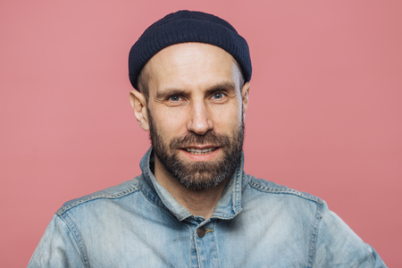 Satisfied male model with thick beard and mustache looks with confident expression into camera, wears fashionable hat and jacket, isolated over pink background. People, emotions, facial expressionsの写真素材