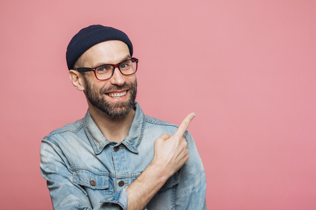 Advertisement concept. Cheerful young bearded man with glad expression wears eyewear, denim jacket and hat, indicates at copy space for your advertisment or promotional text, has happy look.の写真素材