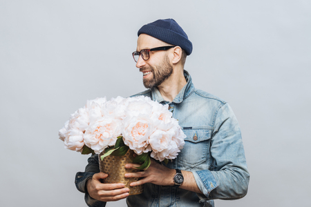Horizontal shot of happy bearded male looks away while holds bunch of white flowers, being in good mood as has date with his girlfriend, makes present for her, isolated over white background.の写真素材