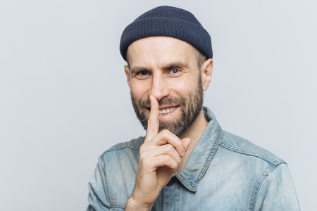 Cheerful attractive male with blue eyes shows silence sign, has satisfied expression, wears fashionable hat and denim jacket, isolated over grey background. Handsome man shows hush sign. Be quiet!の写真素材
