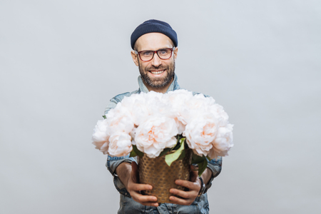 These flowers are for you! Glad smiling bearded male stretches hands while holds bunch of white beautiful flowers, makes present for someone, isolated over studio background, has date with girlfriendの写真素材