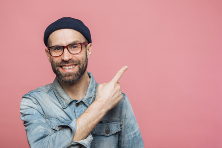 Horizontal shot of cheerful bearded man with happy expression, dressed in fashionable clothing, wears glasses, hat and denim shirt, indicated with fore finger at blank copy space for your advertismentの写真素材