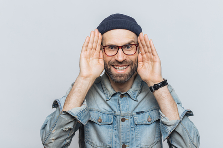 Portrait of happy hipster guy with thick beard and mustache wears stylish hat and glasses, keeps hands near face, tries to see something in darkness, isolated over grey background, has fun indoorの写真素材
