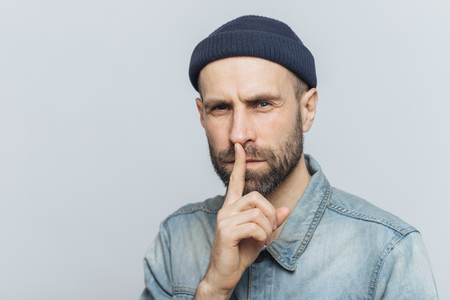Serious pleasant looking bearded male with mysterious expression keeps fore finger on lips, asks to keep personal information confidential, wears stylish hat and denim shirt, isolated on grey wallの写真素材