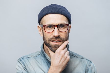 Confident satisfied bearded male keeps hand on chin, listens attentively interlocutor, wears fashionable clothing, isolated over white studio background. People and facial expressions conceptの写真素材
