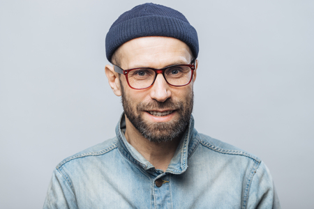 Indoor shot of handsome unshaven male with satisfied expression, rejoices achieving success at work, looks happily through spectacles, isolated over white background. Smiling bearded man indoorの写真素材
