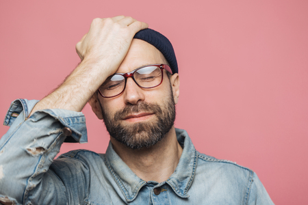 Portrait of depressed middle aged unshaven male closes eyes and keeps hand on forehead, has unhappy expression, isolated over pink background. Fatigue hipster guy has headache after hard workの写真素材