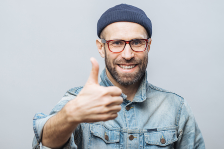 Happy delighted man with stubble raises thumb as shows his approval, dressed in stylish clothing, isolated over white studio background. Satisfied middle aged male gestures indoor. Body languageの写真素材