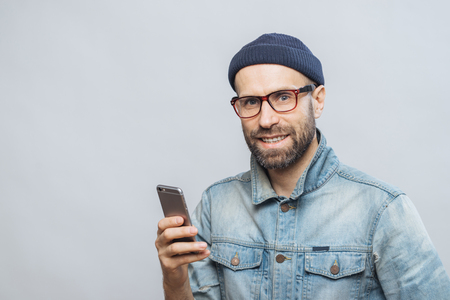 Stusio shot of happy bearded man with stubble holds modern smart phone in hands, enjoys online communication, connected to wireless internet, isolated over white background. Communication conceptの写真素材
