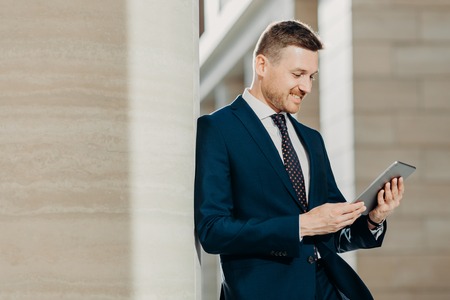 Successful male office worker in black suit uses modern tablet computer for distance work, connected to wireless internet, poses in modern interior, concentrated into screen. Technology conceptの写真素材