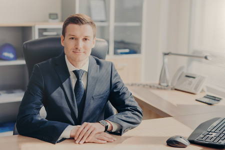Confident company owner in expensive elegant suit sits at work place against office interior, thinks on creation new business plan, has appealing look listens interlocutor attentively. Business peopleの写真素材